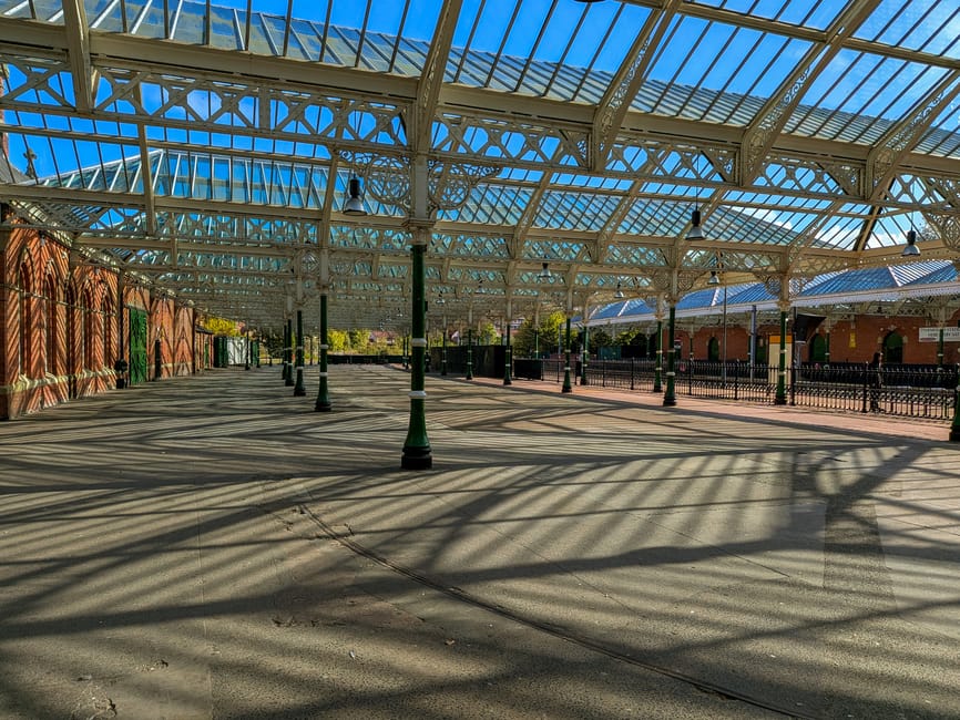 A tall, ornate, reddish-brown wrought iron gate featuring decorative scrollwork, crest accents, and lantern-style light fixtures at the top of the supporting columns, marking the entrance to Queens Ga