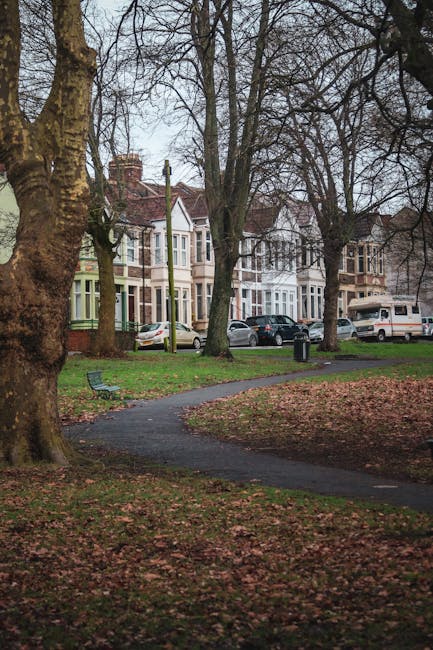 A wide view of a suburban street scene during daytime, featuring a park with a curved paved pathway in the foreground, surrounded by leafless trees and fallen autumn leaves covering the ground. In the background, there are several Victorian-style terraced houses with bay windows, painted in pastel shades, and parked cars along the street, including a white van and smaller vehicles. A few benches are placed near the pathway within the park area. The scene appears to be part of a house removal process, with professional movers from [COMPANY_NAME] possibly preparing to load furniture or moving boxes into their vehicle, which is not visible in this shot. The environment is well-lit with natural daylight, creating clear visibility of the residential surroundings and the nearby street, emphasizing the logistics involved in home relocation or furniture transport in the Queens Park area.